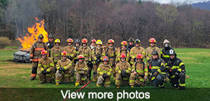 Group photo of firefighter trainees in field with burning car in background. View more photos.
