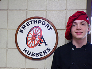 Joshua Raught standing in front of Smethport Hubbers school district logo sign on wall